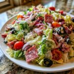 A vibrant, close-up view of a generously dressed grinder salad featuring lettuce, salami, red onion, tomatoes, and olives.