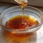 A stream of liquid, likely honey or oil, drips from a spoon into a small glass bowl containing the ingredients for Copycat Apple Cider Vinaigrette.