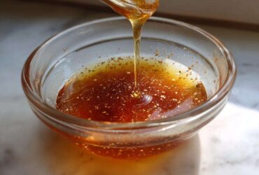 A stream of liquid, likely honey or oil, drips from a spoon into a small glass bowl containing the ingredients for Copycat Apple Cider Vinaigrette.