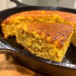 Close-up of a moist slice of golden cornbread with visible corn kernels, freshly baked in a black cast iron skillet.
