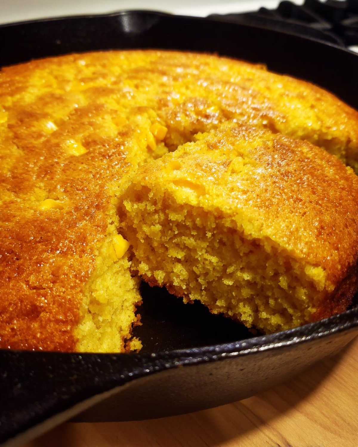 Close-up of a golden brown, moist cornbread with a slice cut out, baked in a black cast-iron skillet.