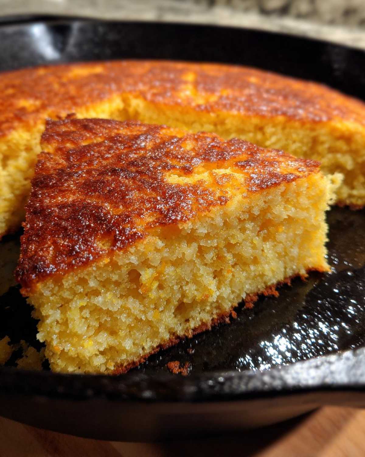 Close-up of a moist slice of golden cornbread with a dark, caramelized crust, served in a black cast iron skillet.