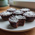 A plate of No-Bake Chocolate Coconut Protein Cups topped with shredded coconut, one cup has a bite taken out showing the dense interior.