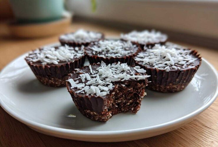 A plate of No-Bake Chocolate Coconut Protein Cups topped with shredded coconut, one cup has a bite taken out showing the dense interior.