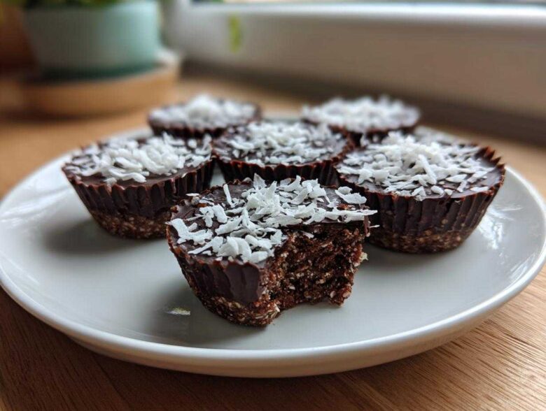 A plate of No-Bake Chocolate Coconut Protein Cups topped with shredded coconut, one cup has a bite taken out showing the dense interior.