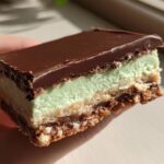 A hand holding a thick slice of No-Bake Chocolate Mint Icebox Bars showing the chocolate top, mint layer, and crumb base.