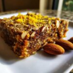 A close-up of a dense No-Bake Lemon Almond Date Bars slice topped with lemon zest and sliced almonds, with whole almonds on the side.