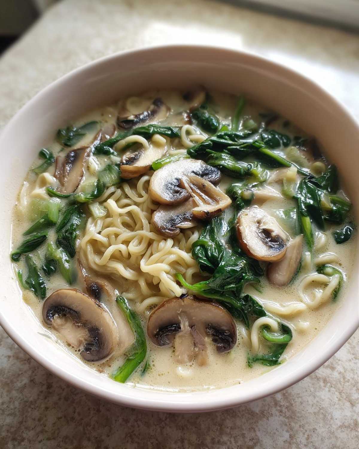 A close-up view of a bowl of One-Pot Creamy Mushroom Spinach Ramen with noodles, sliced mushrooms, and wilted spinach in a creamy broth.