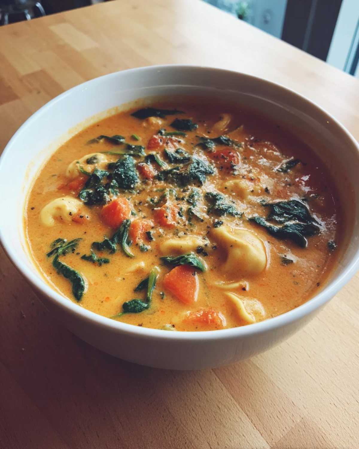 A close-up of a white bowl filled with One-Pot Creamy Tortellini Minestrone soup, featuring tortellini, spinach, and carrots.