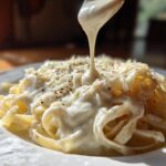 A close-up shot of thick, rich cream sauce being poured over a mound of fettuccine pasta topped with grated cheese.