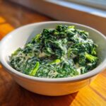 A white bowl filled with vibrant green, rich creamed spinach, highlighted by natural sunlight.