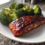 A perfectly cooked fillet of Sheet Pan Honey Harissa Salmon topped with a dark, glossy glaze, served next to bright green steamed broccoli florets.