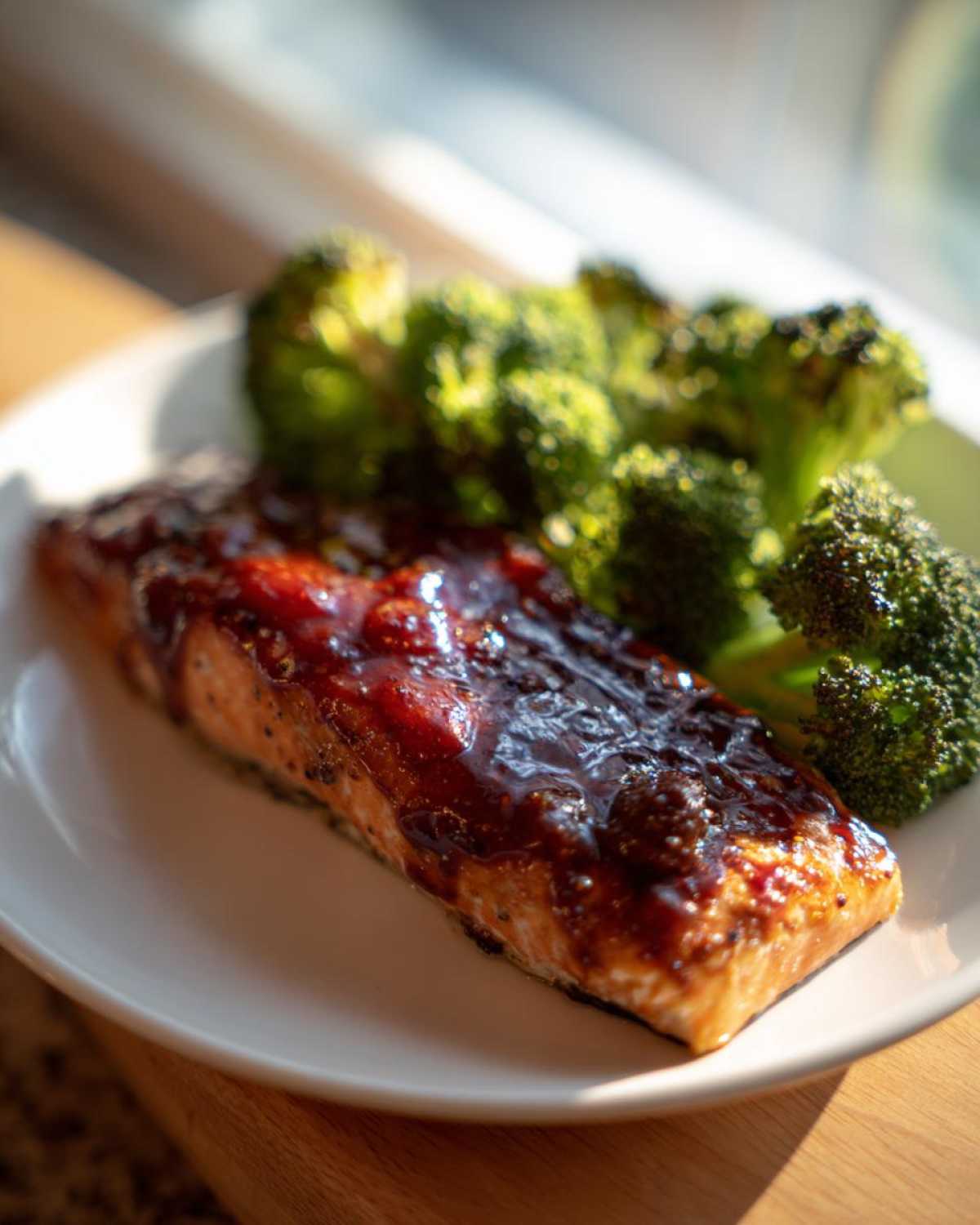 A close-up of a glazed salmon fillet topped with a dark, shiny sauce, served next to roasted broccoli on a white plate.