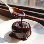 A spoonful of rich, silky chocolate ganache is being poured over a small, round dessert on a white plate.