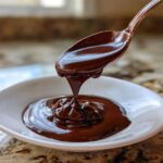 A spoonful of rich, glossy chocolate ganache drips back into a small white bowl.