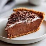 A decadent slice of french silk pie featuring a thick chocolate filling, whipped cream, and dark chocolate shavings on a graham cracker crust.