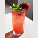 A tall glass filled with bright pink Sparkling Strawberry Basil Lemonade, garnished with a fresh strawberry and basil sprig.