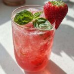 A tall glass filled with bright pink Sparkling Strawberry Basil Lemonade, garnished with fresh basil leaves and a whole strawberry.