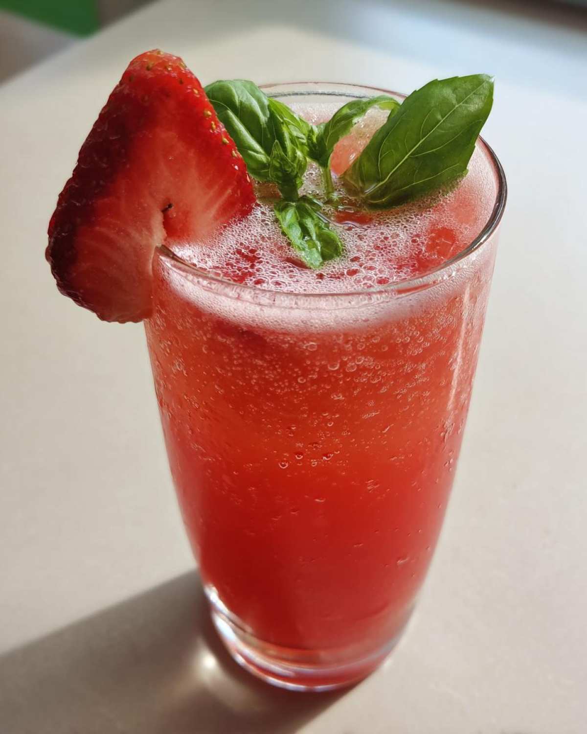 Close-up of a tall glass filled with bright red Sparkling Strawberry Basil Lemonade, garnished with a strawberry slice and fresh basil leaves.