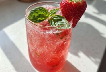 A tall glass filled with bright pink Sparkling Strawberry Basil Lemonade, garnished with fresh basil leaves and a whole strawberry.
