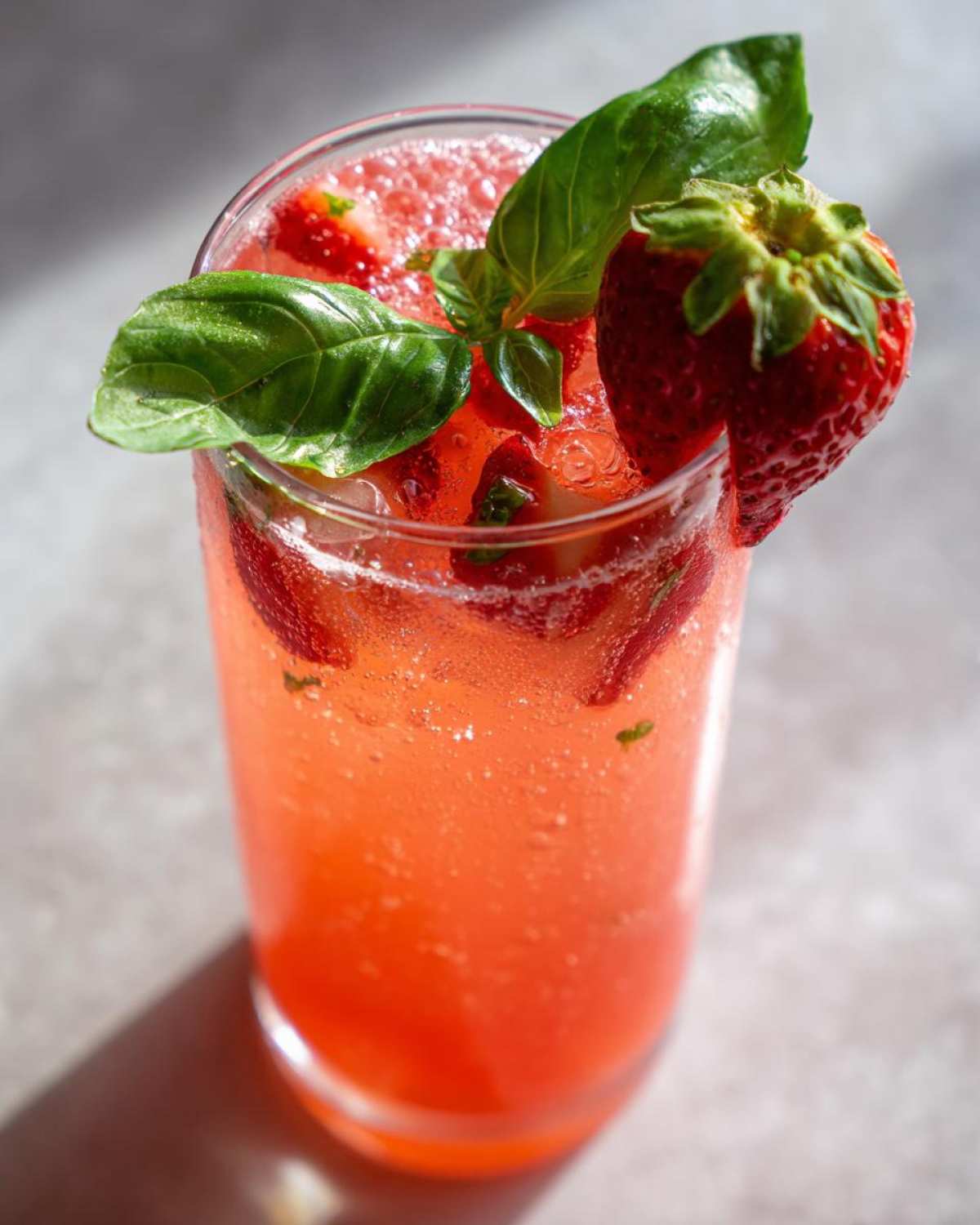 Close-up of a tall glass filled with vibrant Sparkling Strawberry Basil Lemonade, garnished with fresh basil leaves and a whole strawberry.