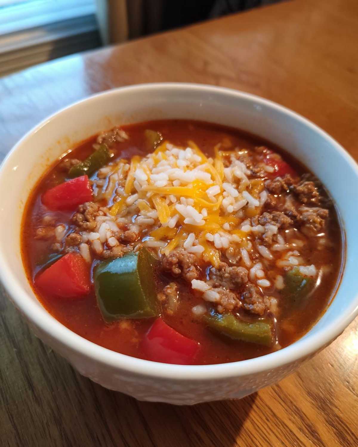 A close-up of a white bowl filled with stuffed pepper soup, featuring ground beef, red and green peppers, rice, and shredded cheese.