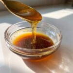 Close-up of a golden liquid, likely honey or maple syrup, being drizzled from a spoon into a small glass bowl containing the base for Copycat Apple Cider Vinaigrette.