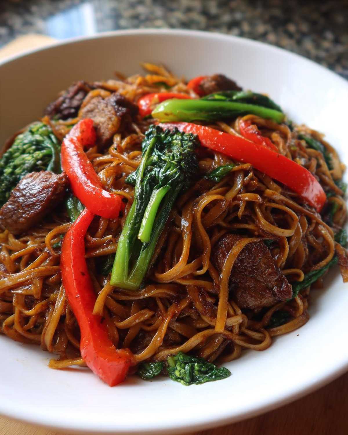 A close-up of a serving of savory Thai drunken noodles mixed with beef, red bell peppers, and green leafy vegetables in a white bowl.