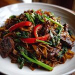 A close-up, appetizing shot of glossy, dark brown Thai drunken noodles mixed with beef, bright red bell peppers, and green Chinese broccoli.