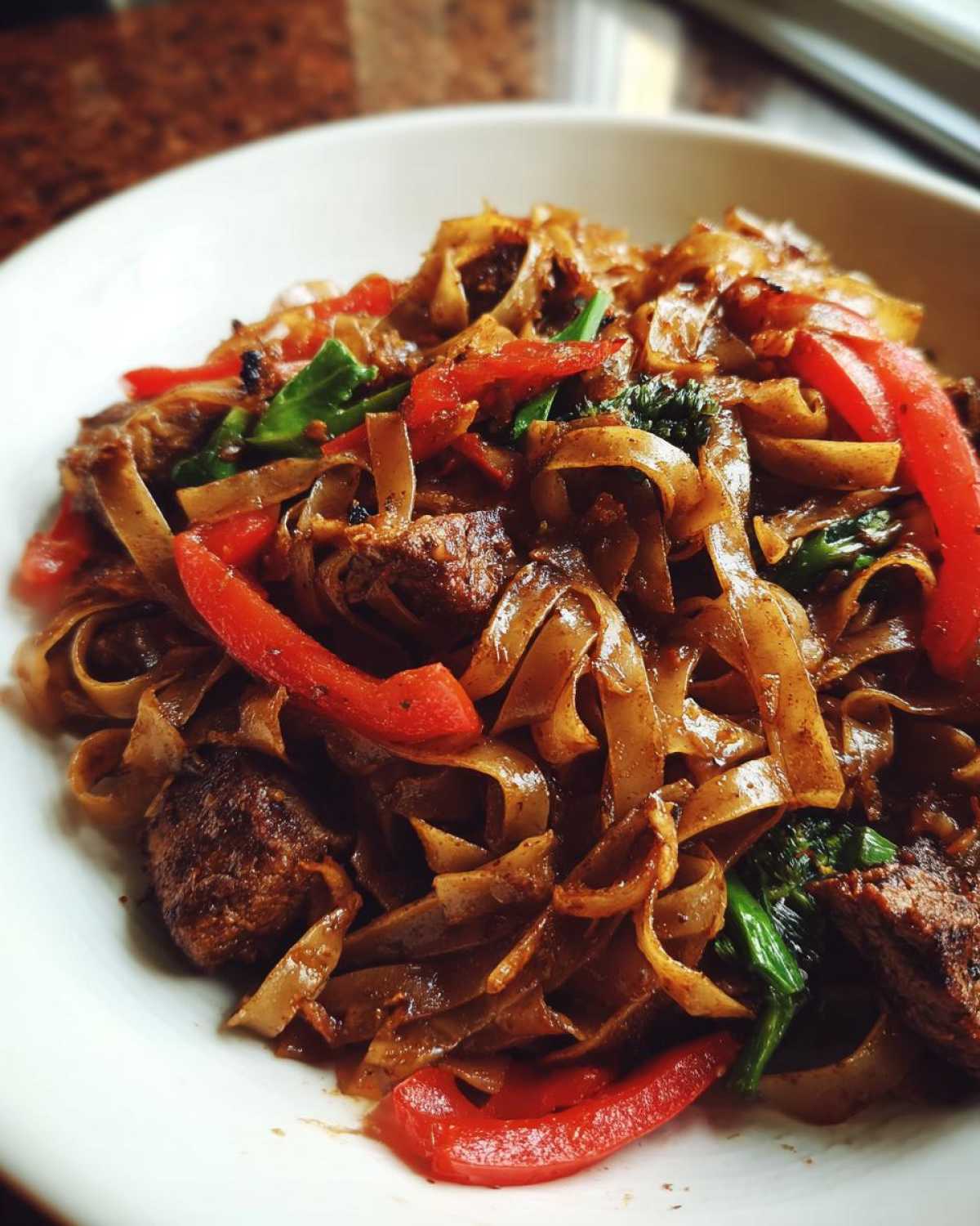 A close-up shot of a white bowl filled with glossy, dark brown Thai drunken noodles, mixed with pieces of beef and bright red bell peppers.