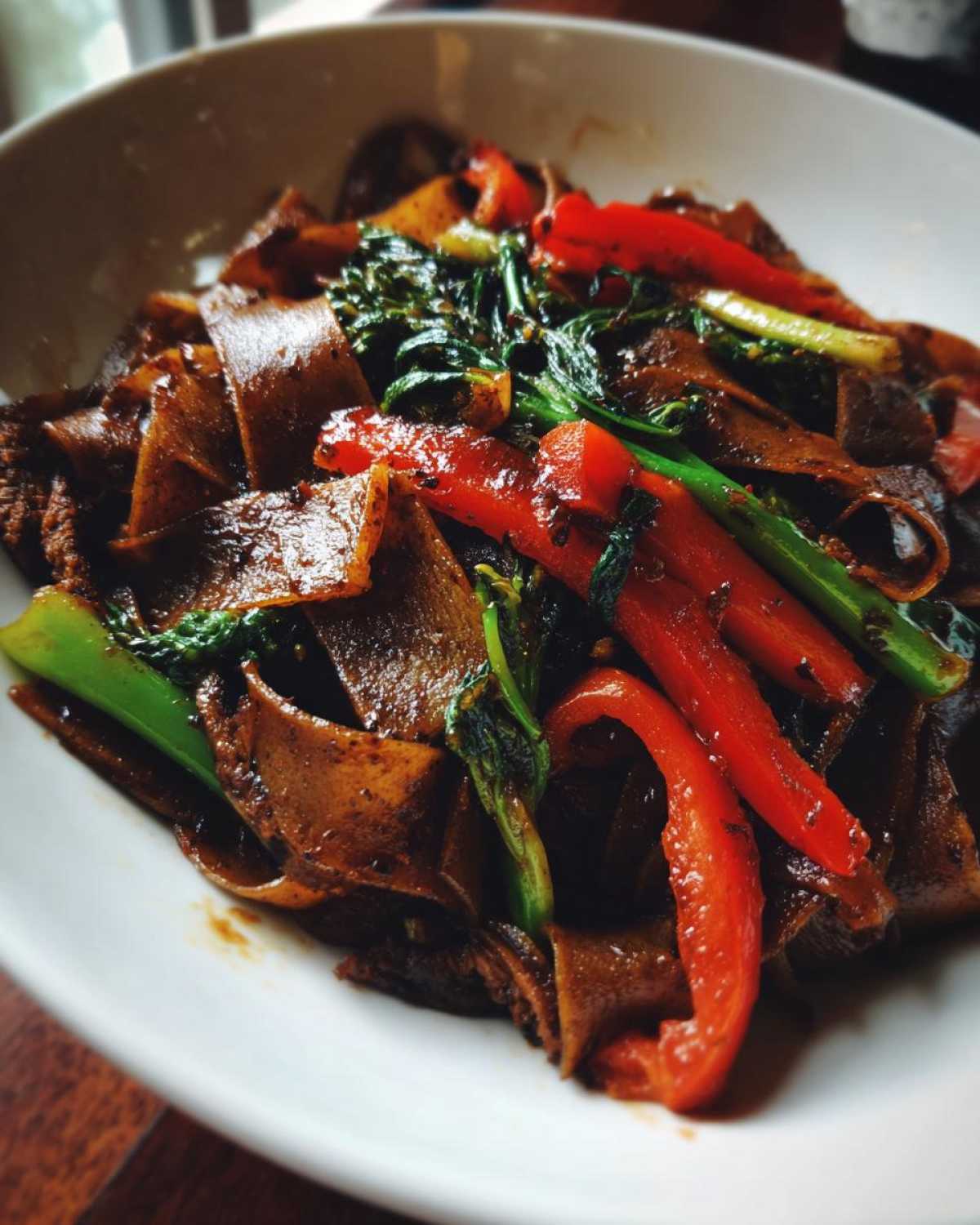 A close-up serving of glossy, dark brown Thai drunken noodles mixed with bright red bell peppers and green Chinese broccoli.