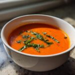 Close-up of a white bowl filled with bright orange tomato basil soup, garnished with fresh basil leaves.