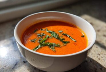 Close-up of a white bowl filled with bright orange tomato basil soup, garnished with fresh basil leaves.