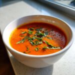 A close-up of bright orange tomato basil soup garnished with fresh, thinly sliced basil leaves.