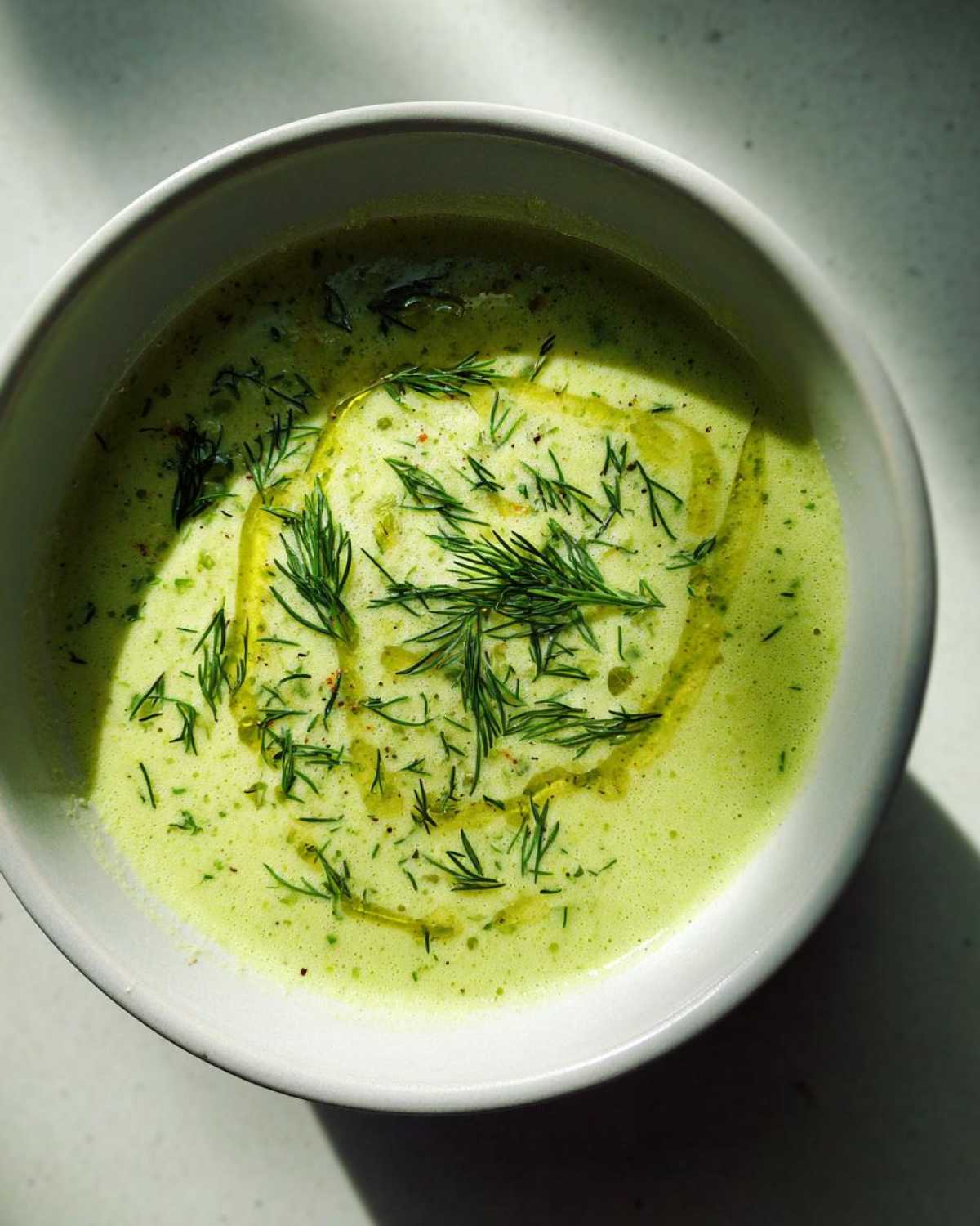 Overhead view of a bowl of bright green Vegan Creamy Cucumber Dill Soup, garnished with fresh dill and olive oil.