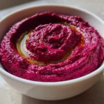 Close-up of vibrant magenta Vegan Creamy Roasted Beet Hummus swirled in a white bowl, topped with olive oil.