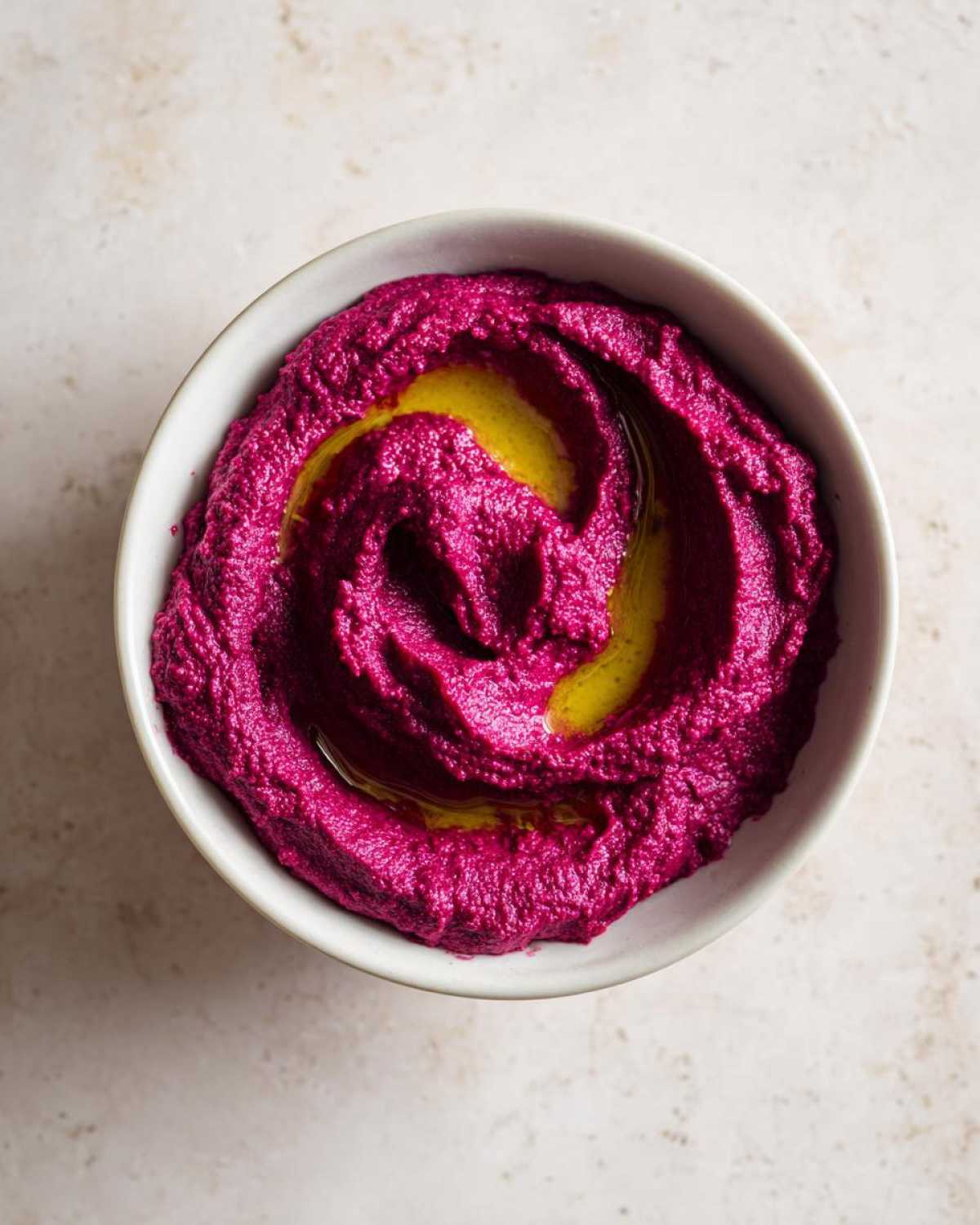 Overhead view of bright magenta Vegan Creamy Roasted Beet Hummus swirled in a white bowl and drizzled with olive oil.