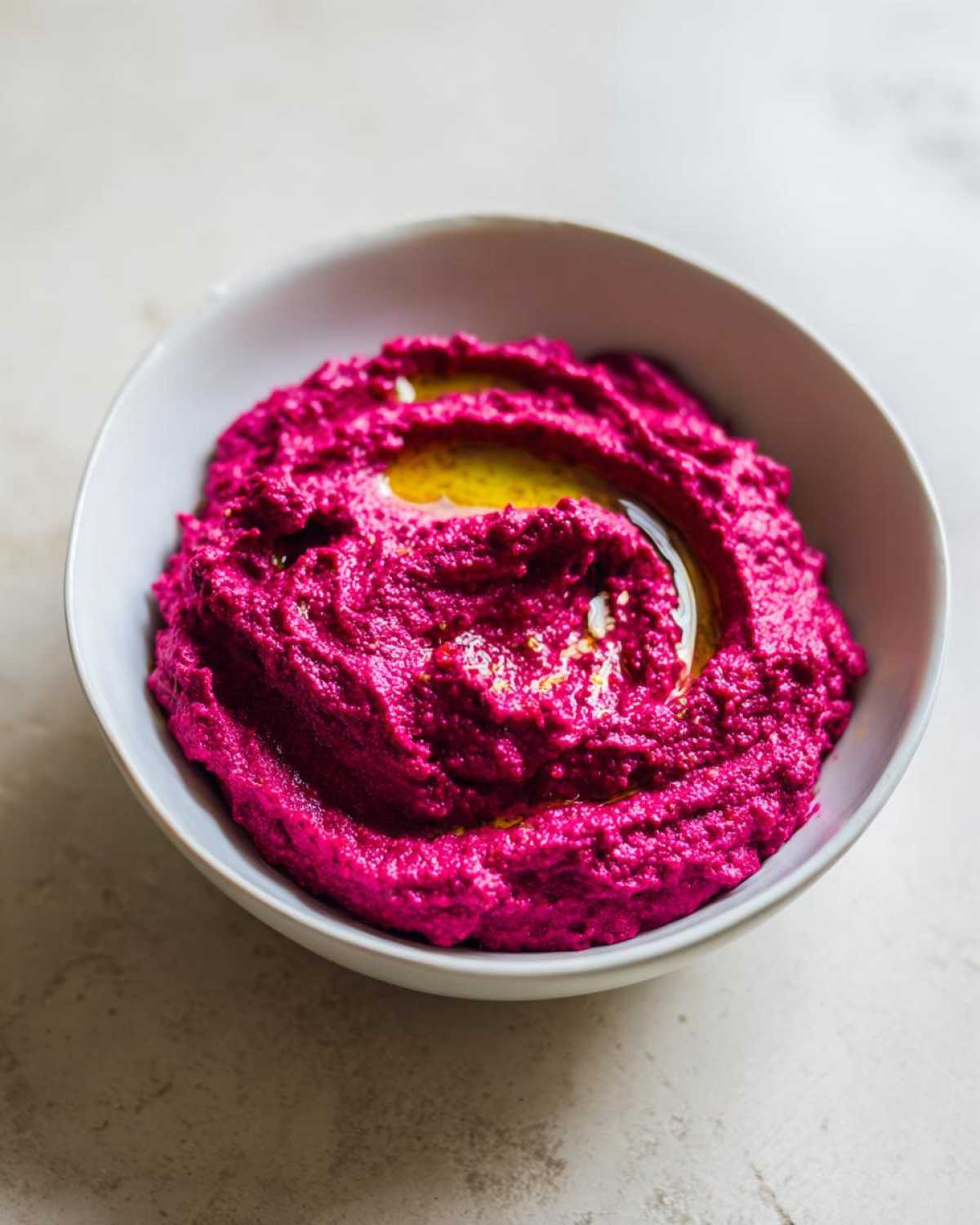 A close-up of vibrant pink Vegan Creamy Roasted Beet Hummus swirled in a white bowl, drizzled with olive oil.