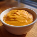 A close-up of a thick, vibrant orange cheese sauce served in a small white bowl on a wooden surface.