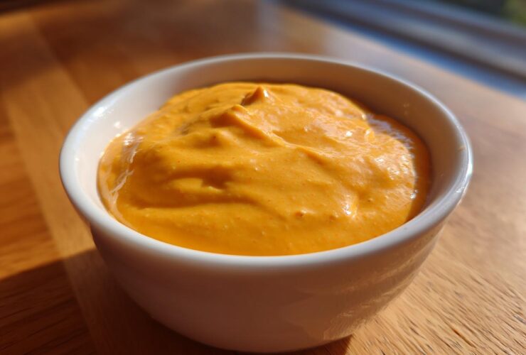 A close-up of a thick, vibrant orange cheese sauce served in a small white bowl on a wooden surface.